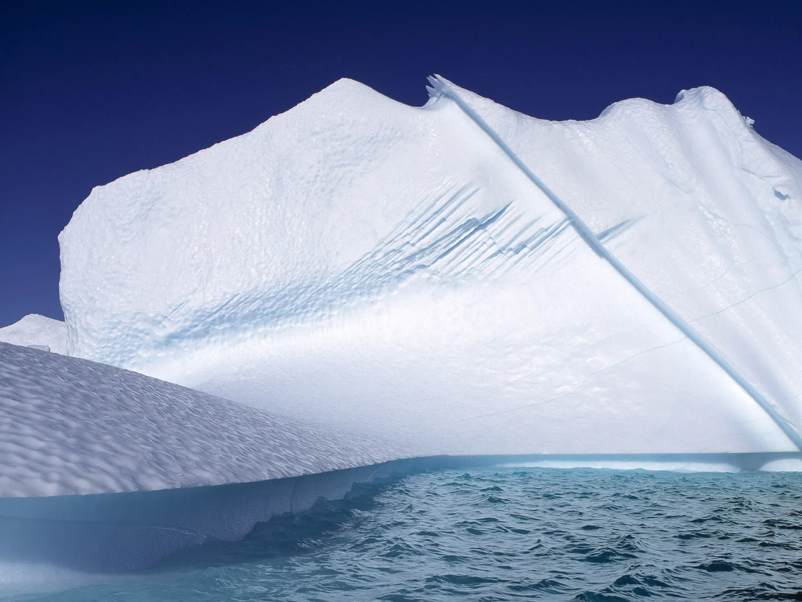 Sunlight illuminates the textured surface of a large iceberg floating on deep blue ocean water.
