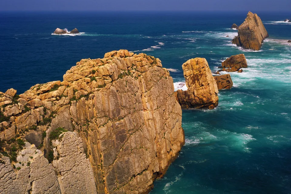 Sunlit jagged cliffs overlook a turquoise ocean with crashing waves.