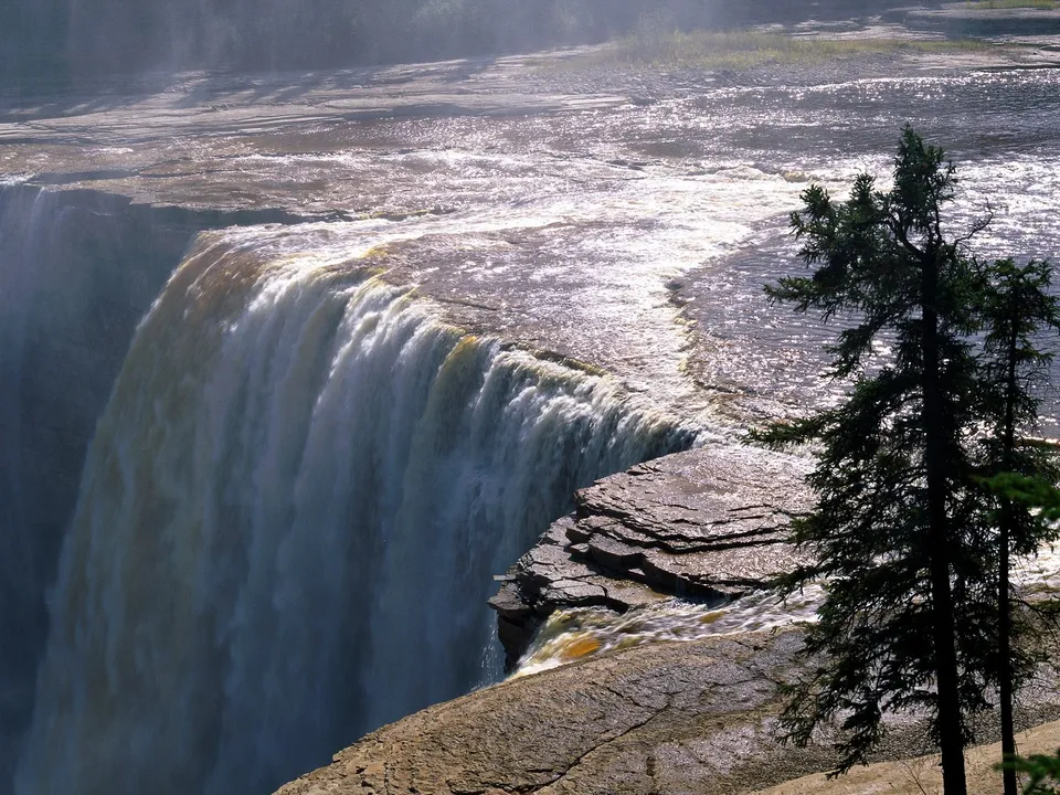 A powerful waterfall plunges over a wide, layered rock ledge in bright sunlight.