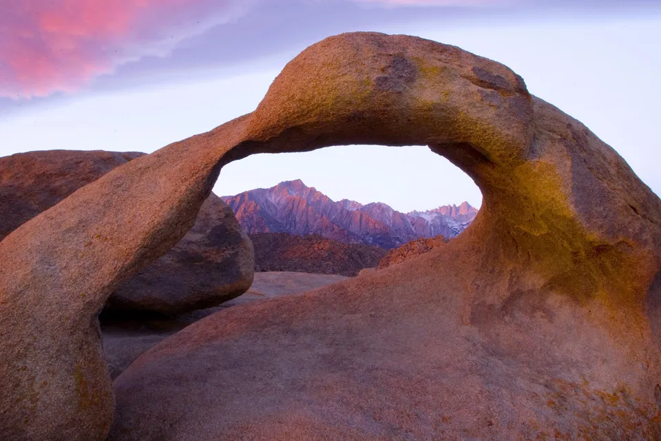 A natural rock arch frames snow-capped mountains bathed in the warm light of sunrise.
