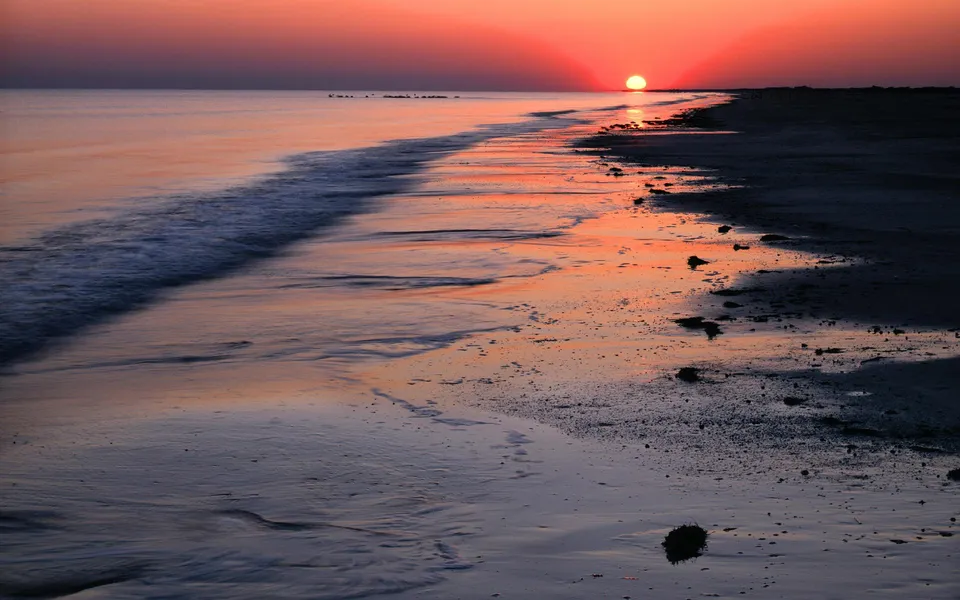 The sun rises over a calm ocean, casting a golden glow on the wet sand and gentle waves.