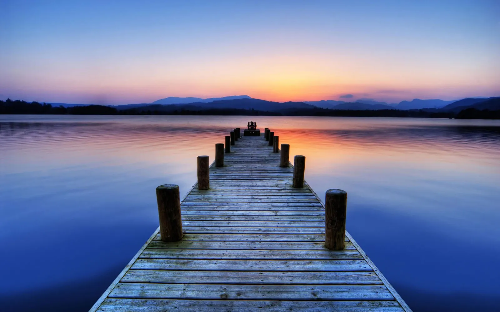 A wooden pier extends into a calm lake during a colorful sunrise, with mountains in the distance.