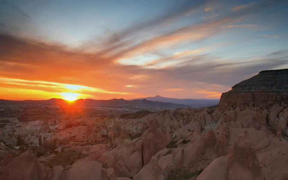 A vibrant sunrise illuminates the unique rock formations and distant mountains of Cappadocia.