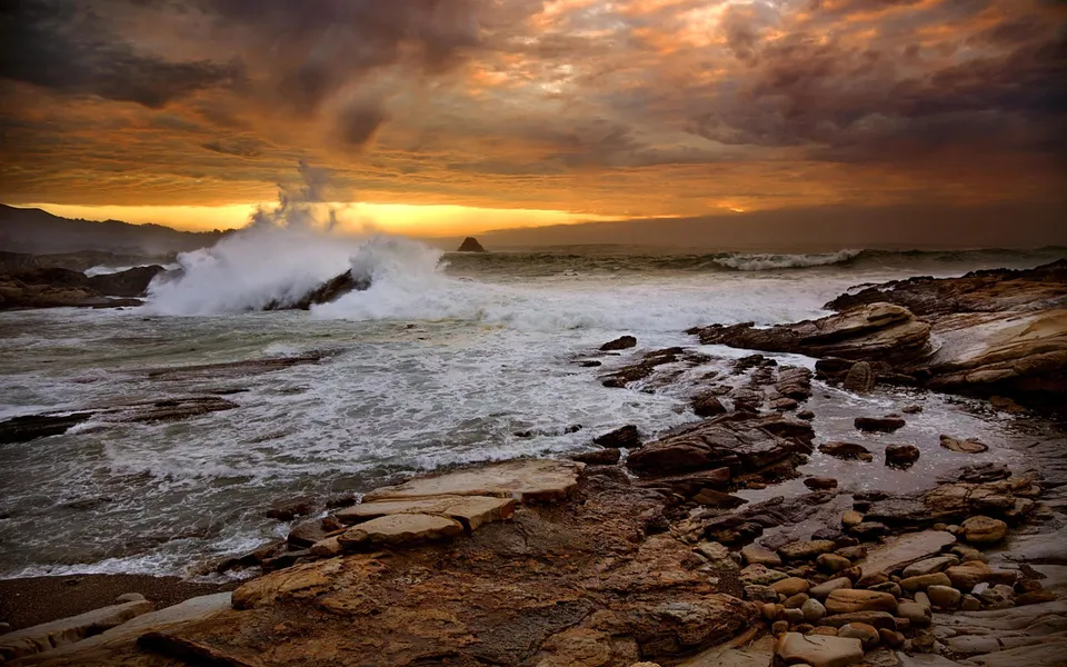 Waves crash against a rocky coastline during a dramatic sunrise with orange and dark clouds.