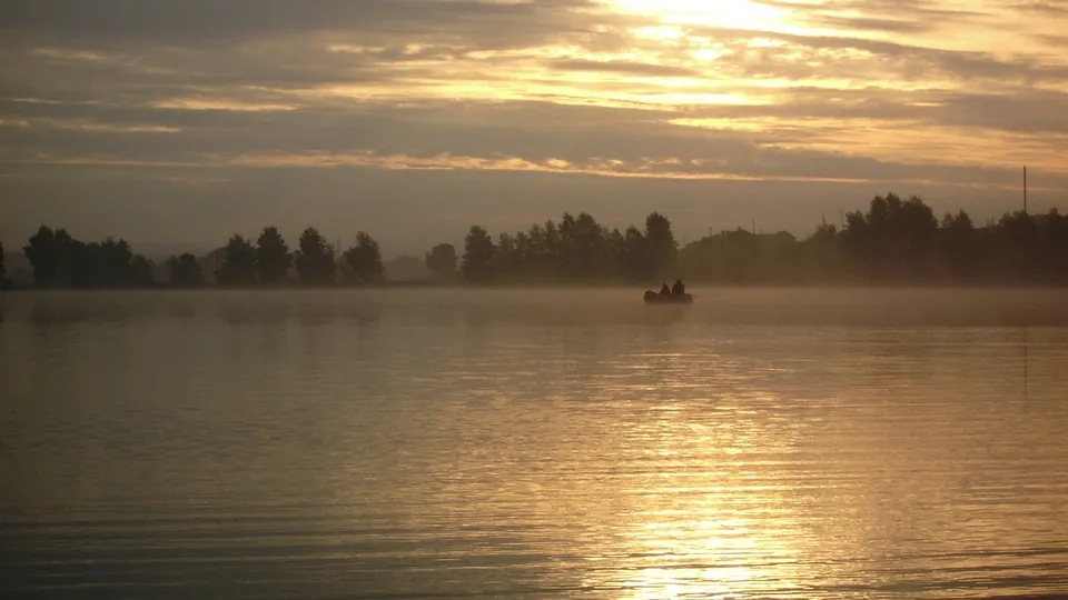 Two people in a small boat fishing on a misty lake at sunrise, with golden light reflecting on the water.