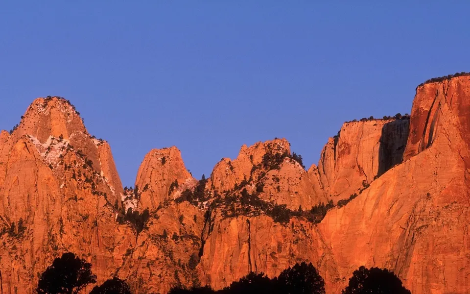The sun casts a golden glow on the rugged peaks of desert mountains under a clear blue sky.