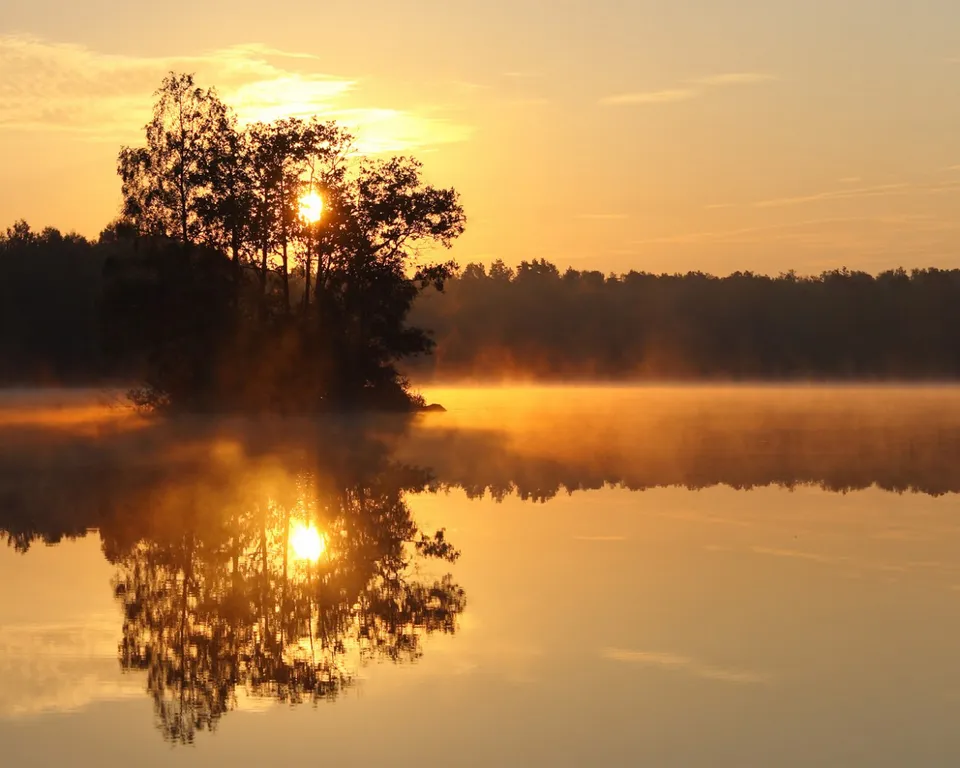 A serene sunrise over a misty lake, with the sun peeking through trees on a small island, casting a reflection on the water.