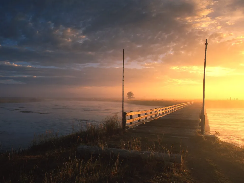 A wooden bridge stretches across a misty river at sunrise, with golden light illuminating the sky.