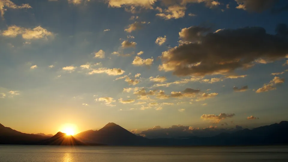 A vibrant sunrise casts golden rays over a calm lake and silhouetted mountains.