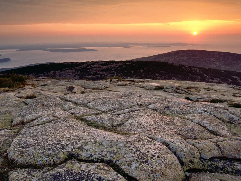 A vibrant sunrise paints the sky with orange and yellow hues over a rocky mountain landscape and a distant ocean.