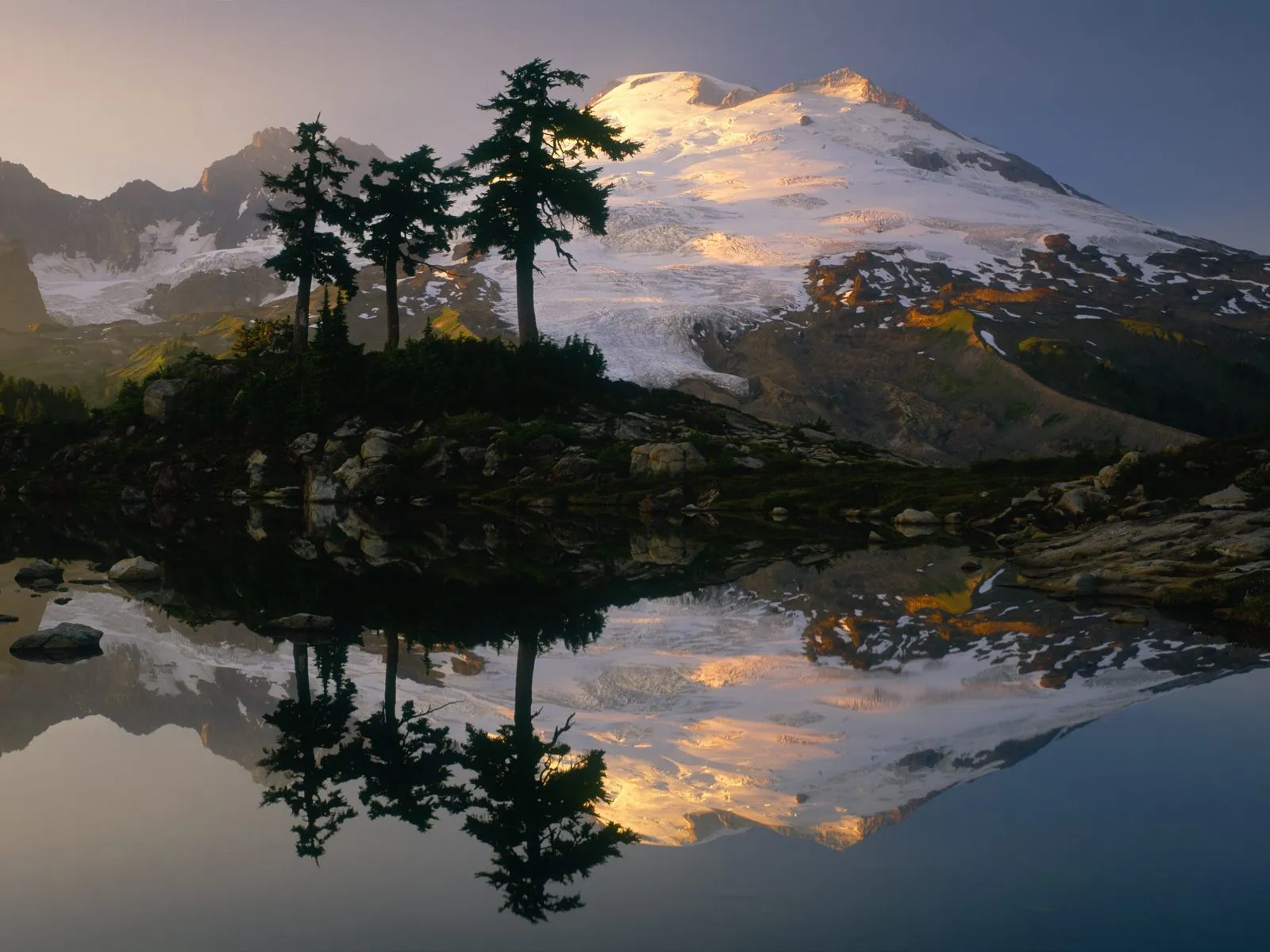 Sunrise illuminates a snow-covered mountain peak, reflected in a calm lake with silhouetted trees.