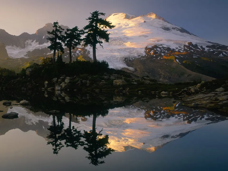 Sunrise illuminates a snow-covered mountain peak, reflected in a calm lake with silhouetted trees.