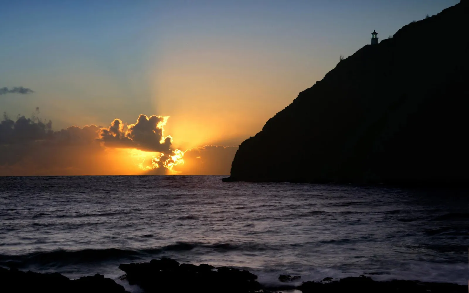 A dramatic sunrise with golden rays breaking through clouds over a dark ocean, with a silhouetted cliff and lighthouse on the right.