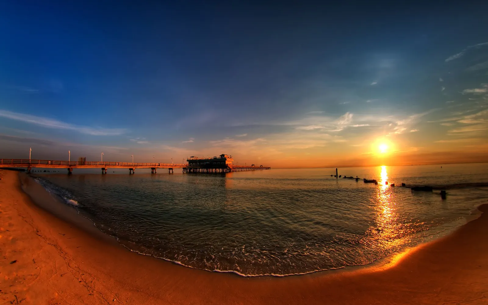 A wide panoramic view of a beach and ocean at sunrise, with a pier extending into the water.