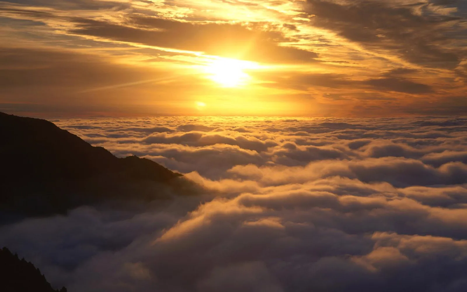 The sun rises over a vast expanse of clouds, with a mountain peak visible on the left.