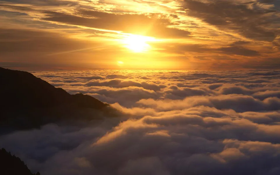 The sun rises over a vast expanse of clouds, with a mountain peak visible on the left.