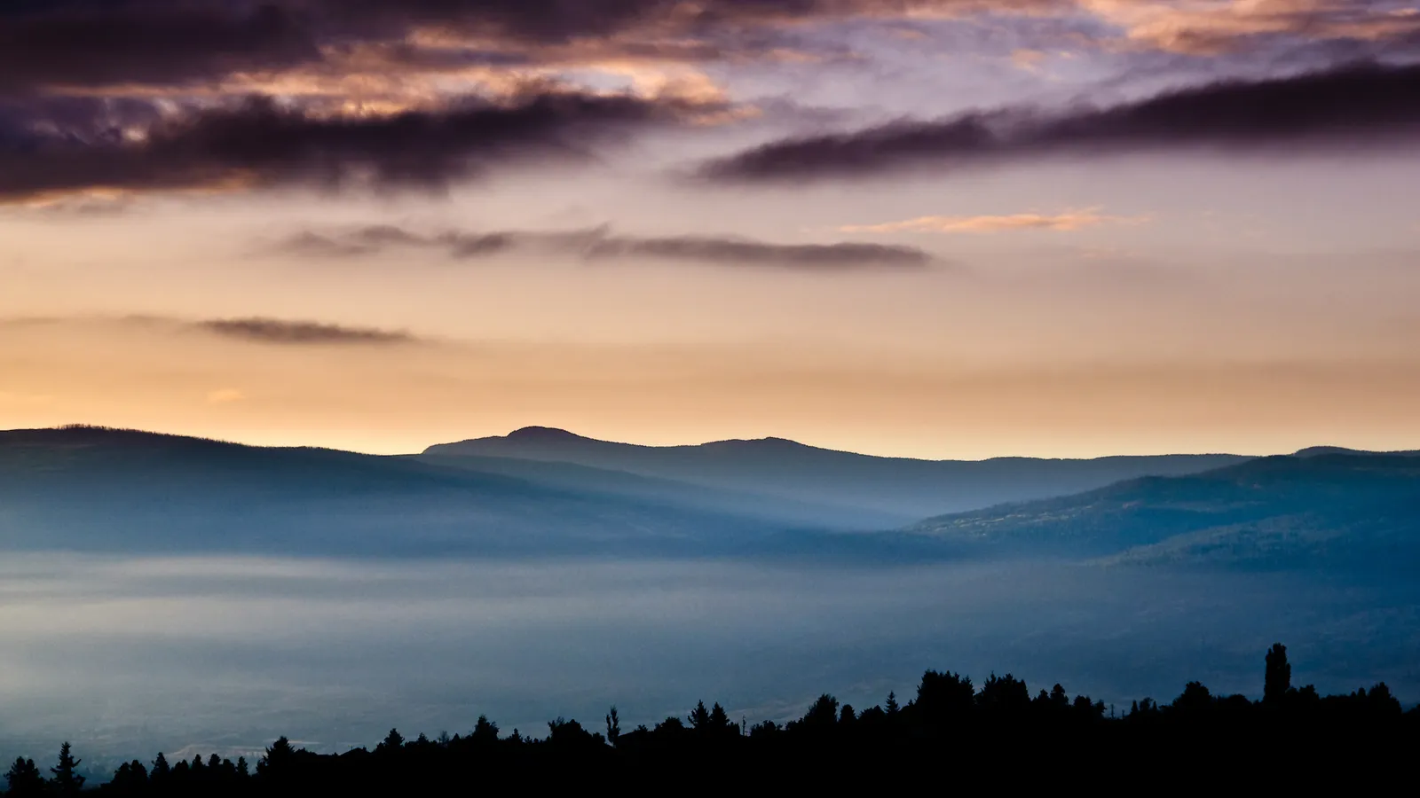 Sunrise paints the sky with warm colors over layers of misty mountains shrouded in blue fog.