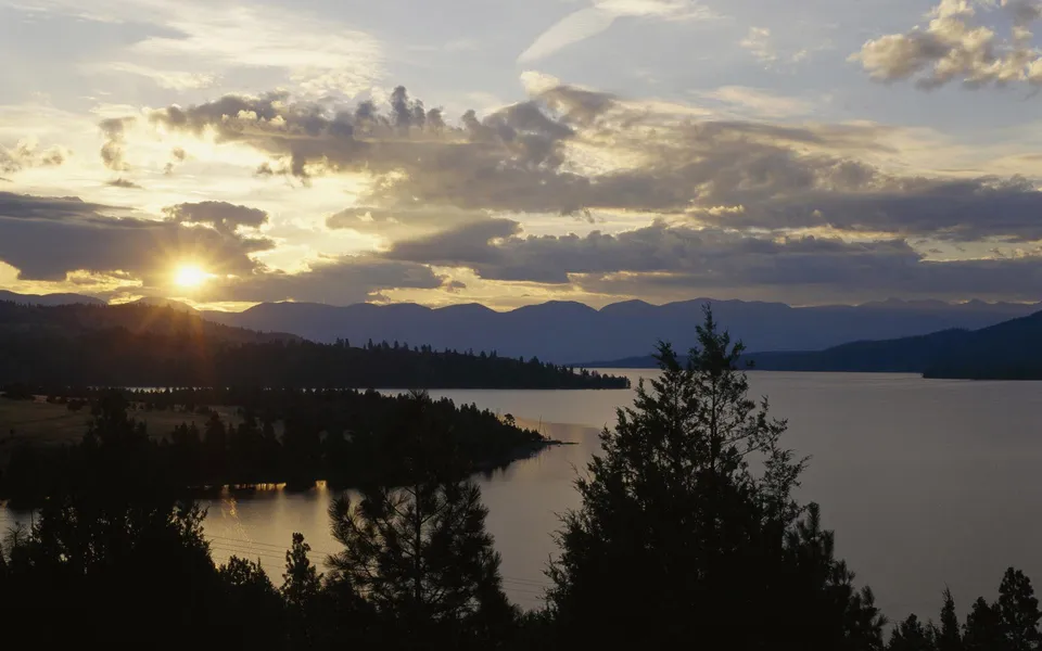 The sun rises over a calm lake, casting golden light through dramatic clouds and illuminating distant mountains.