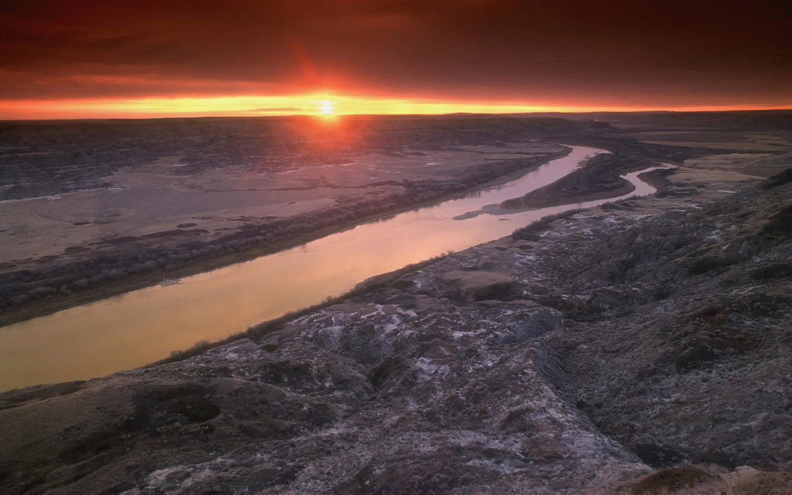 A wide river winds through a valley at sunrise, with the sun low on the horizon and dramatic orange clouds.