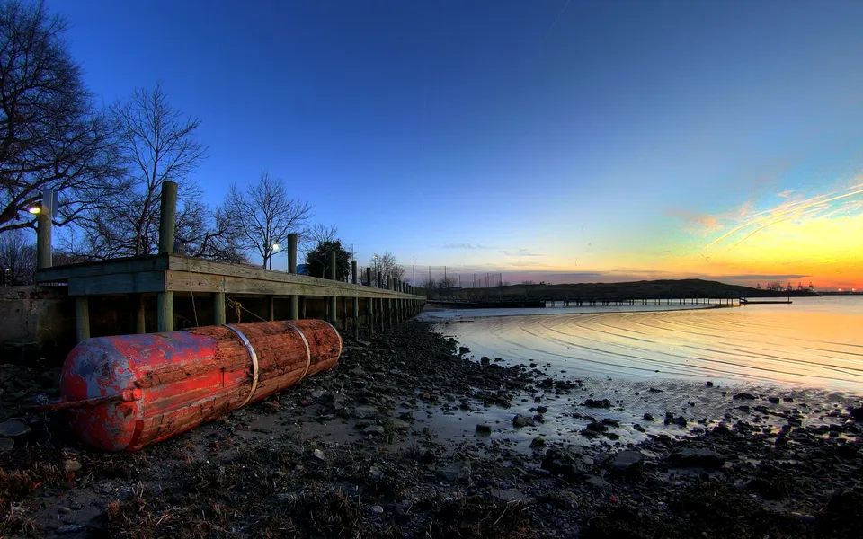 A wooden pier stretches into calm water at sunrise, with a red buoy in the foreground.