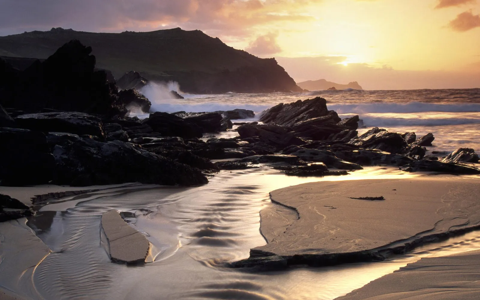 A dramatic sunrise illuminates a rocky coastline with waves crashing and sand patterns.