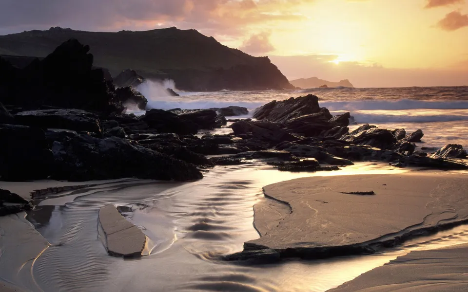 A dramatic sunrise illuminates a rocky coastline with waves crashing and sand patterns.