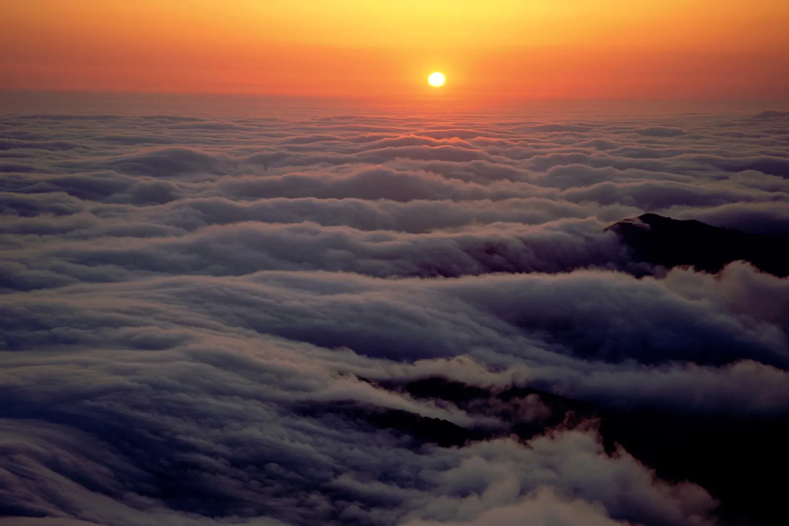 The sun rises over a vast expanse of clouds, with mountain peaks visible below.
