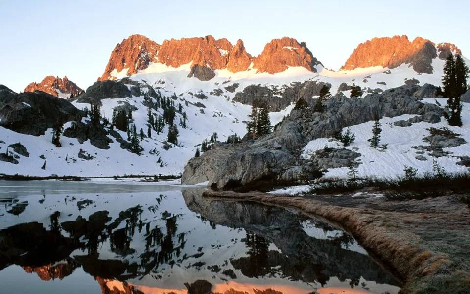 A serene lake reflects snow-capped mountains illuminated by the warm glow of sunrise.