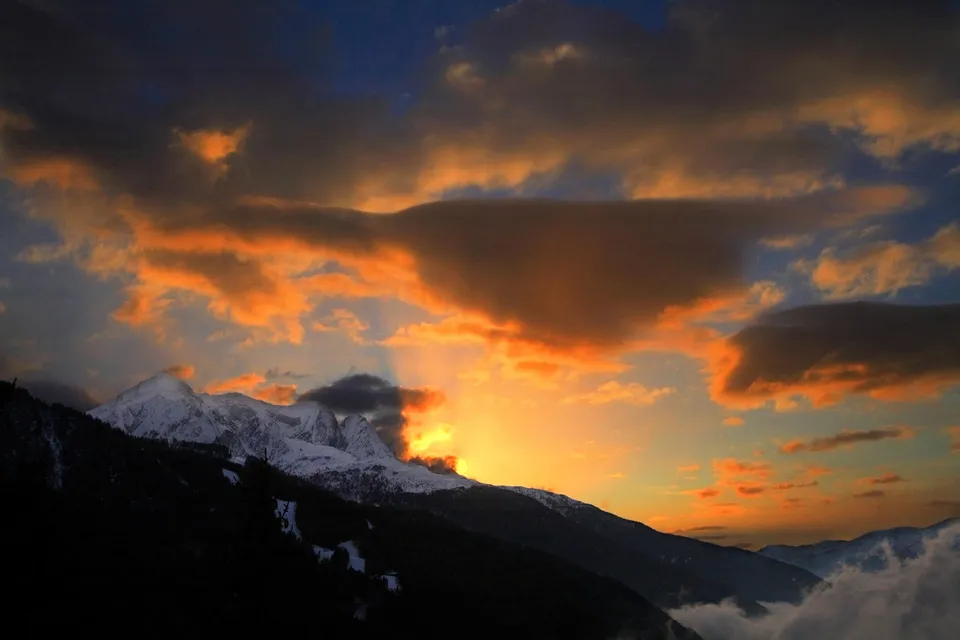 A dramatic sunset with orange and dark clouds over snow-capped mountains.