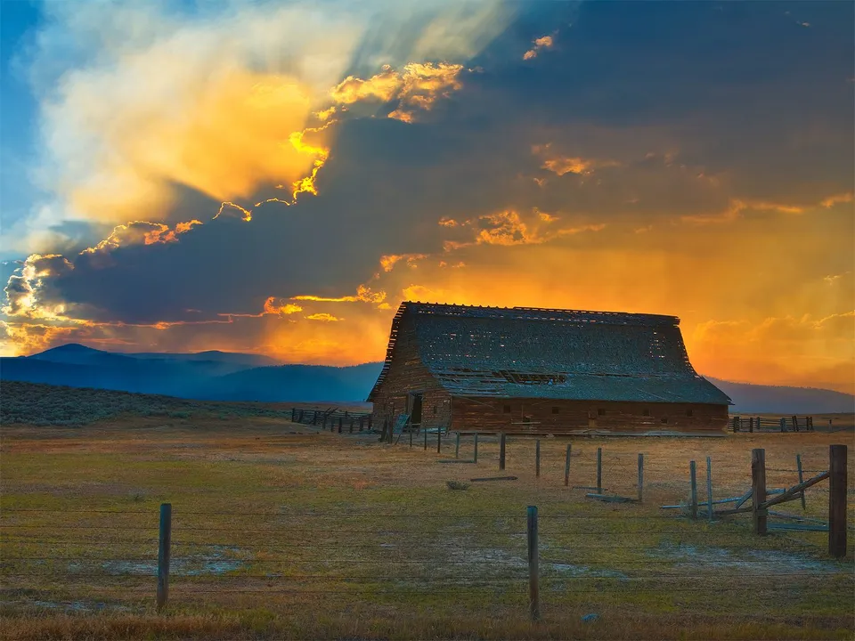 An old wooden barn sits in a field under a dramatic sunset sky with rays of light breaking through the clouds.