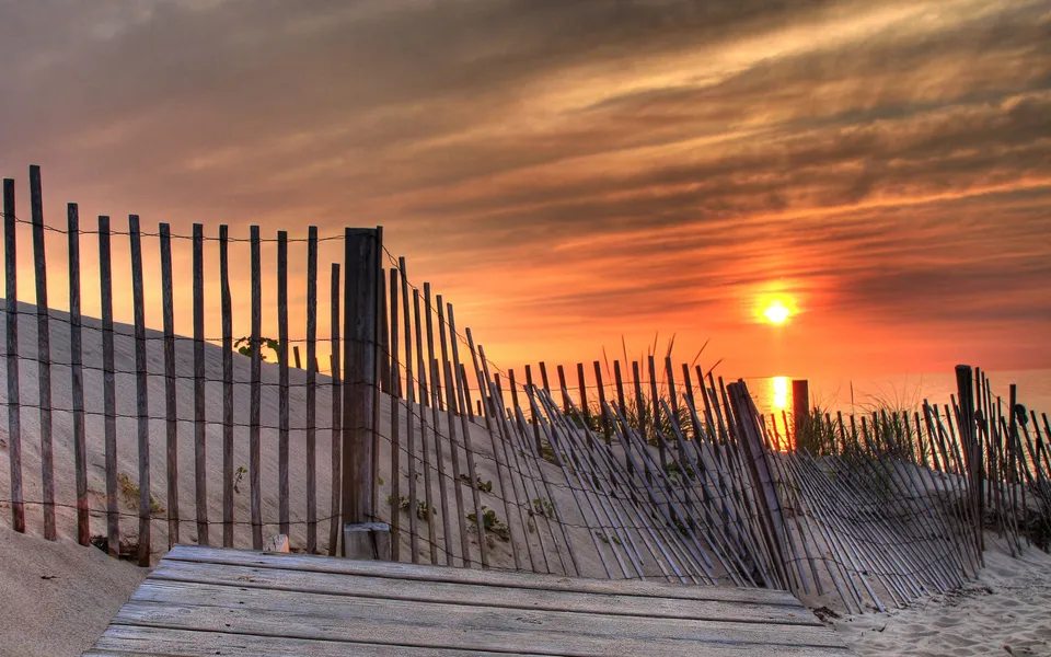 A wooden beach fence with sand dunes and grass in the foreground, silhouetted against a vibrant sunset over the ocean.