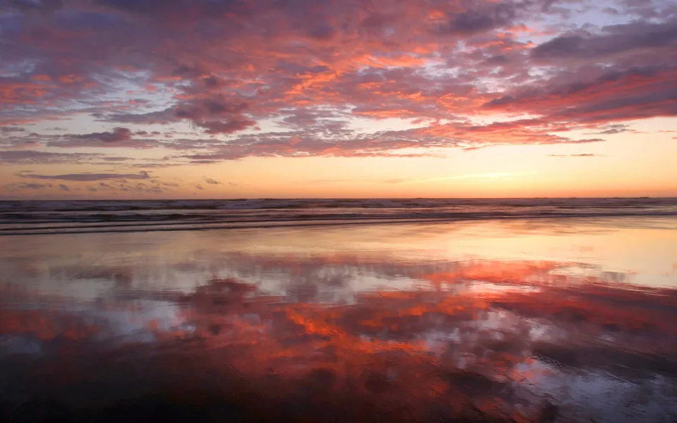 A colorful sunset with pink and orange clouds reflected on the wet sand of a beach.