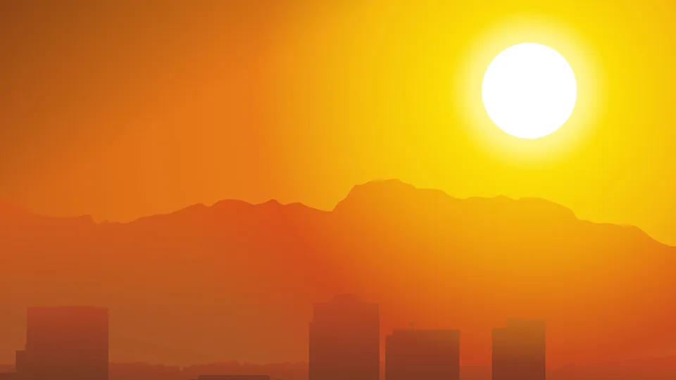 A bright sun sets behind hazy mountains, casting an orange glow over a silhouetted cityscape.