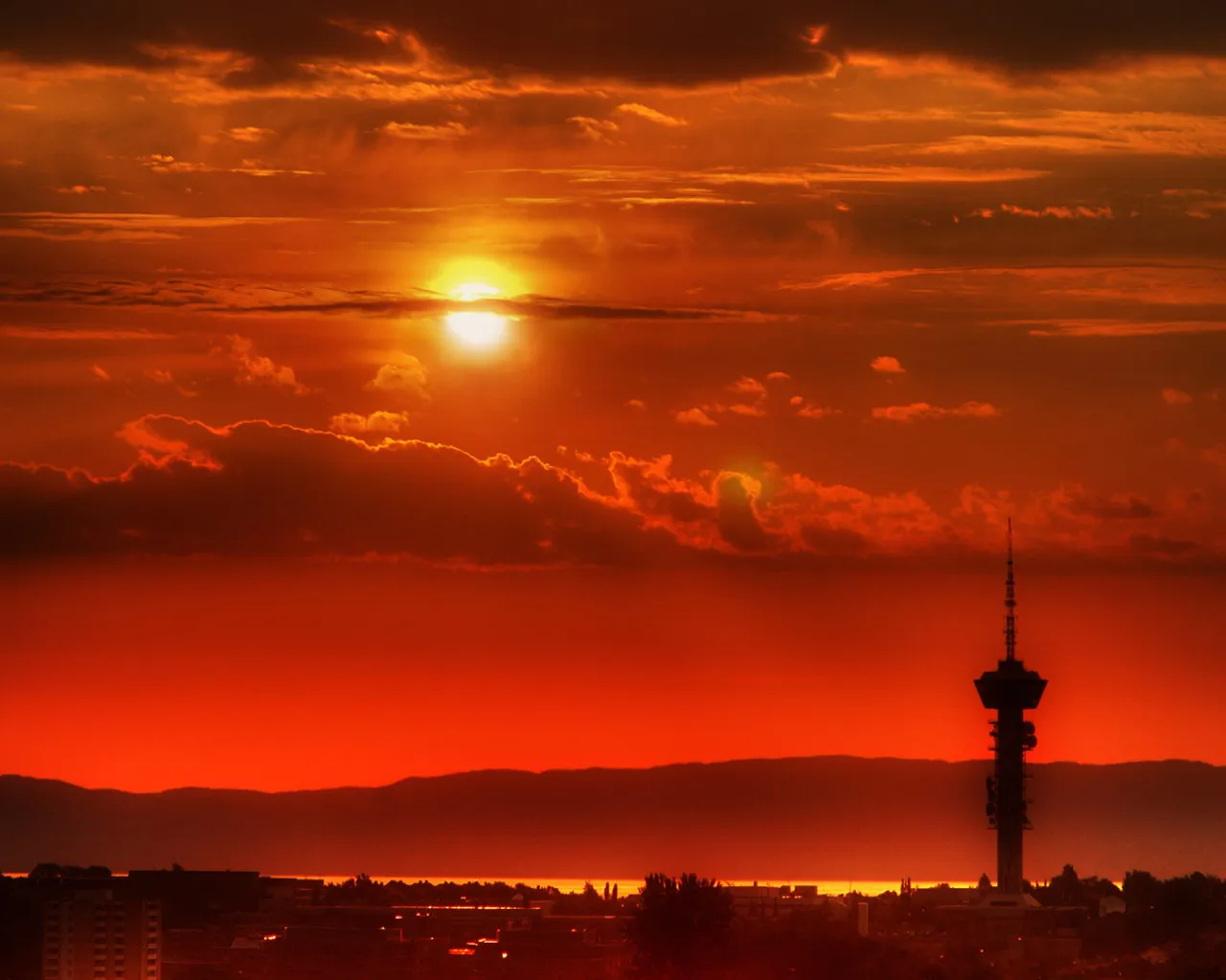 A bright sun sets behind clouds, casting a fiery orange glow over a city skyline with a tall tower.