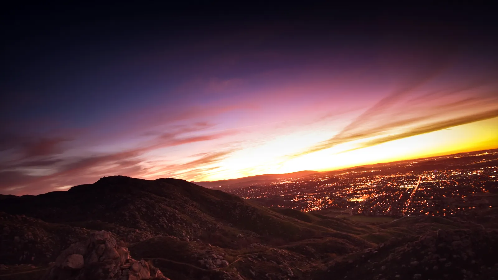 A vibrant sunset with streaks of pink and orange clouds over a sprawling city at dusk, viewed from a dark mountain.