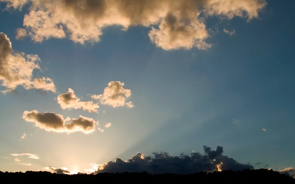Sunbeams break through clouds at sunset, casting golden light over a dark, silhouetted horizon.
