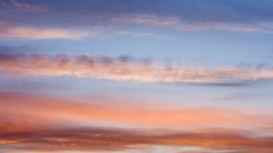 Wispy clouds are illuminated with soft pink, orange, and purple hues against a pale blue sky.