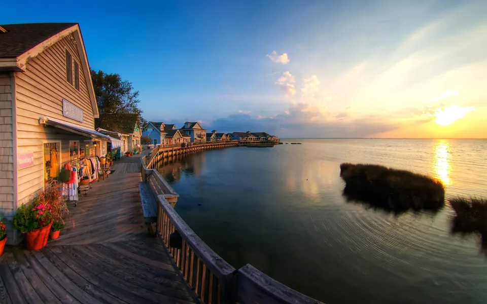 A wooden boardwalk lines shops along a calm body of water at sunset, with the sun low on the horizon.