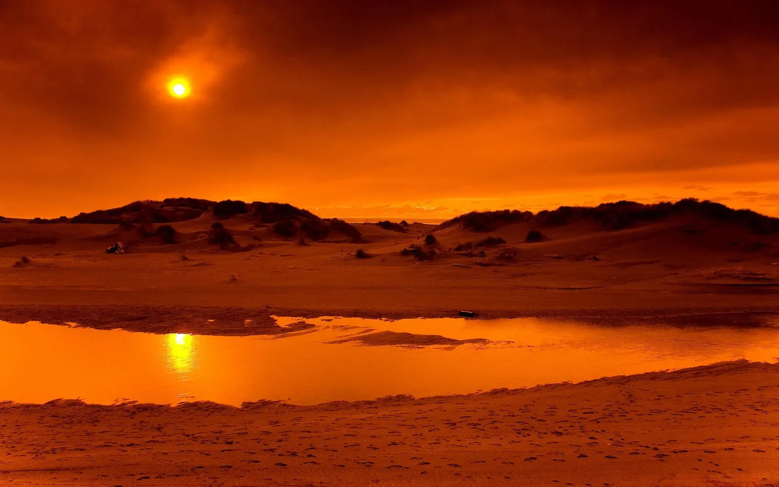 A vibrant orange sunset illuminates coastal sand dunes and a shallow pool of water reflecting the sky.