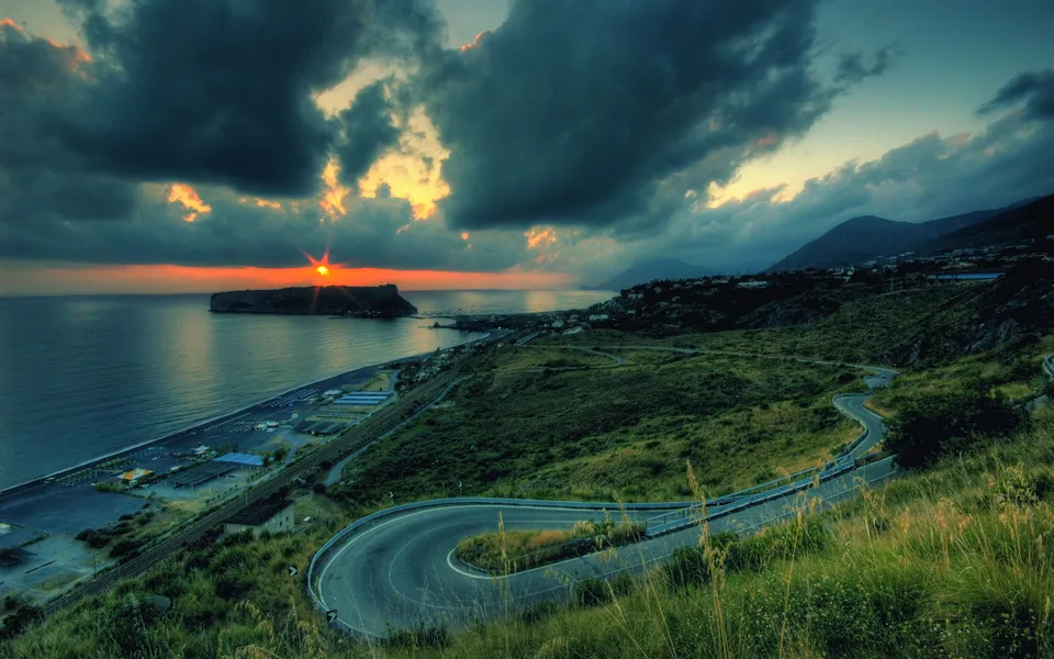 A winding road descends a grassy hill towards a coastline with a dramatic sunset over the sea.