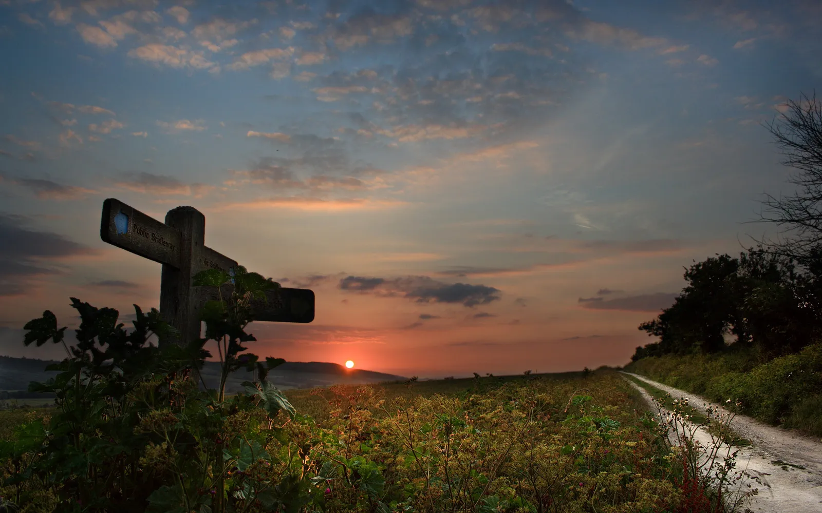 A wooden signpost points down a path towards a sunset over rolling hills.