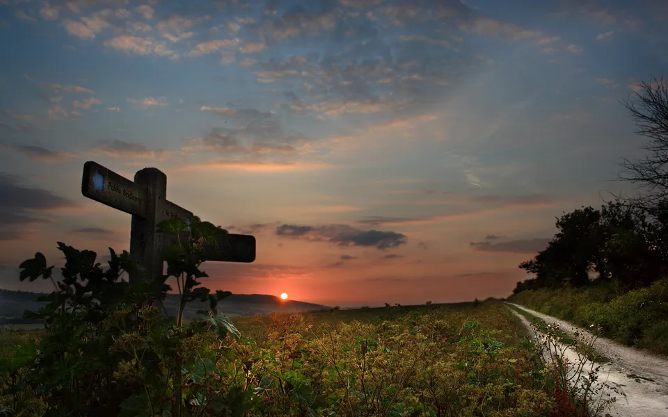 A wooden signpost points down a path towards a sunset over rolling hills.