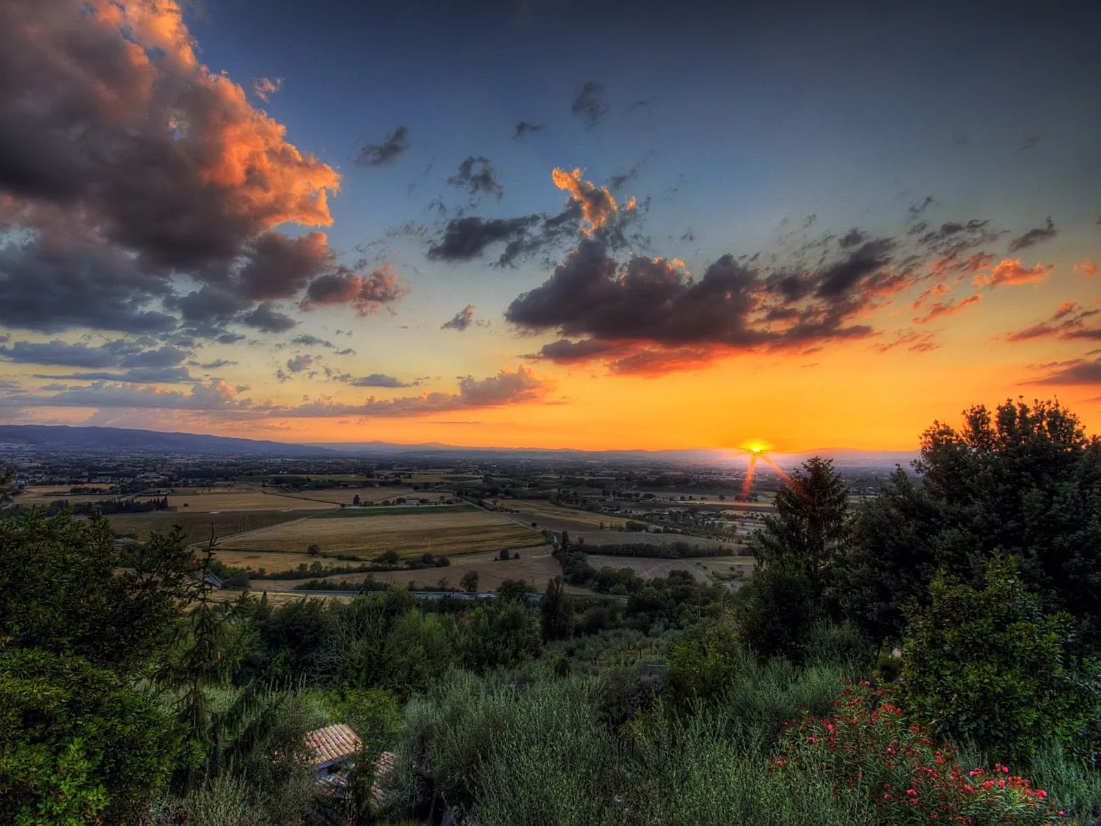 A vibrant sunset paints the sky with orange and pink hues over a vast rural landscape with fields and trees.