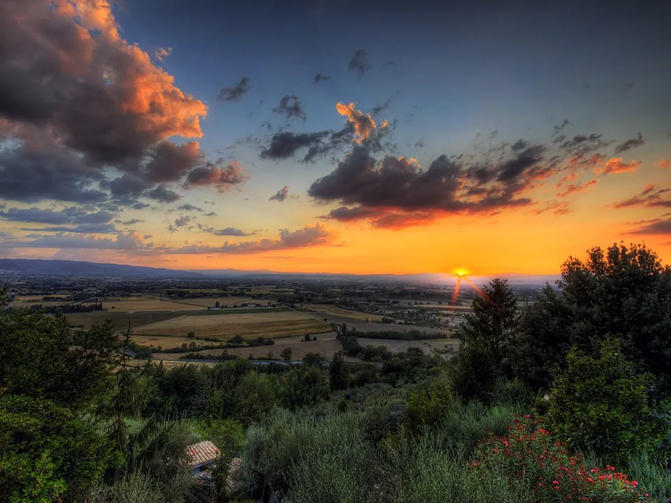 A vibrant sunset paints the sky with orange and pink hues over a vast rural landscape with fields and trees.
