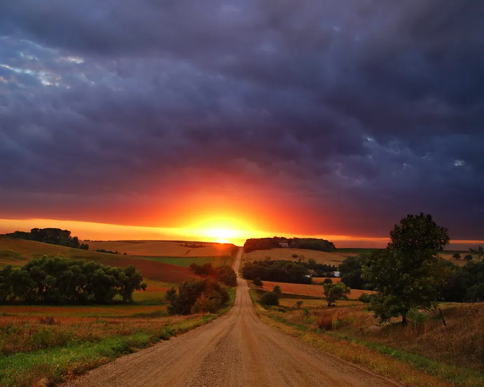 A dirt road leads through rolling hills towards a vibrant sunset with dramatic clouds.