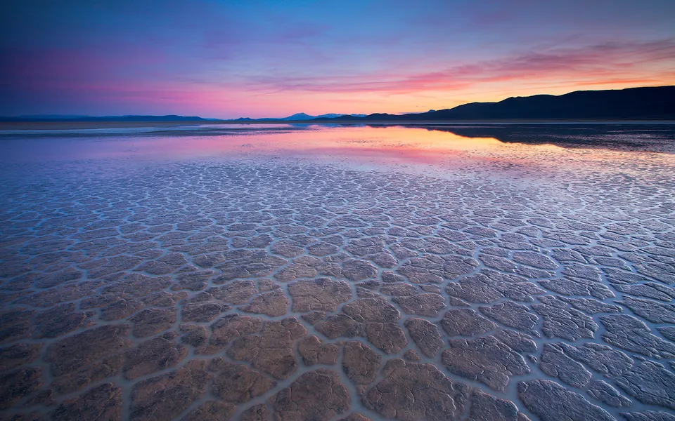 A vibrant sunset with pink and blue hues reflects on a shallow lake covering cracked earth.