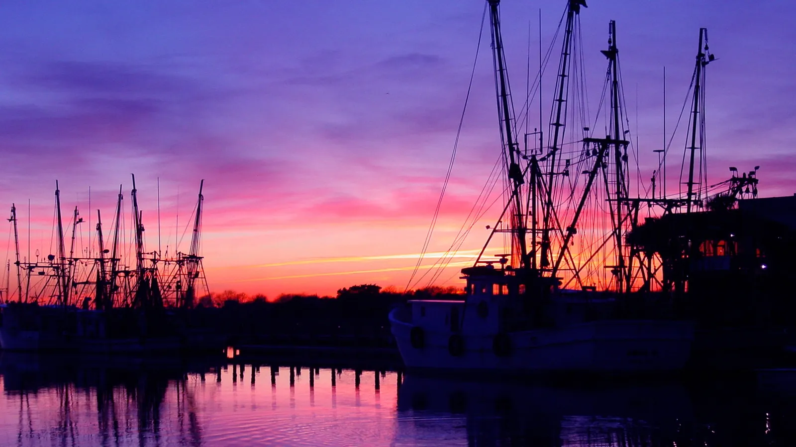 Silhouetted fishing boats in a harbor during a vibrant sunset with purple and pink skies.