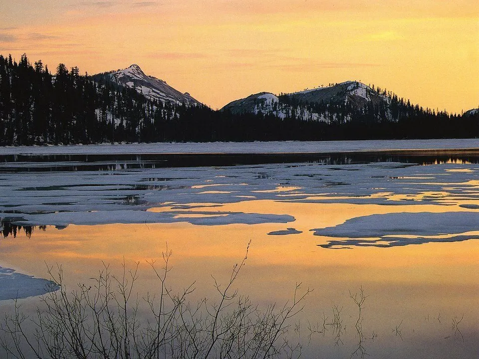 A serene mountain lake at sunset, with ice floes reflecting the warm sky and snow-capped peaks in the background.