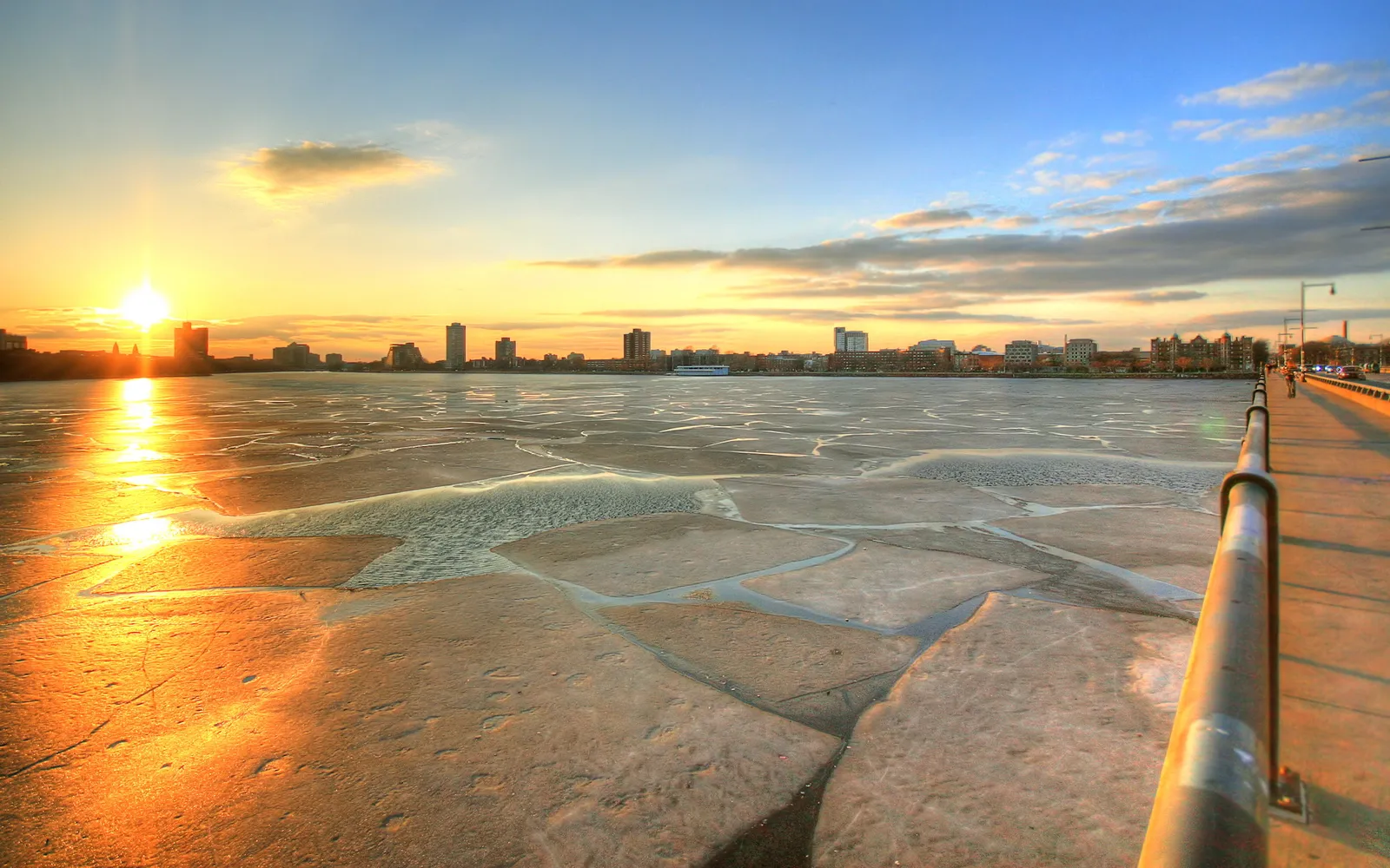 A bright sun sets over a frozen river with a city skyline in the distance and a walkway in the foreground.