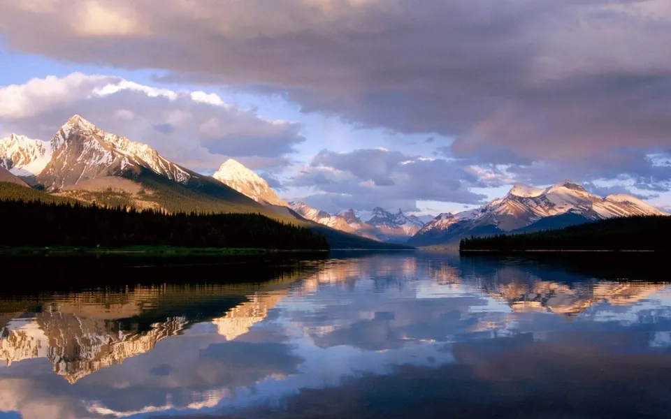 Snow-capped mountains glow with sunset colors reflected in a calm lake.
